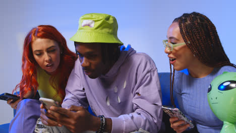 Studio-Shot-Of-Young-Gen-Z-Friends-Sitting-On-Sofa-Sharing-Social-Media-Post-On-Mobile-Phones-1